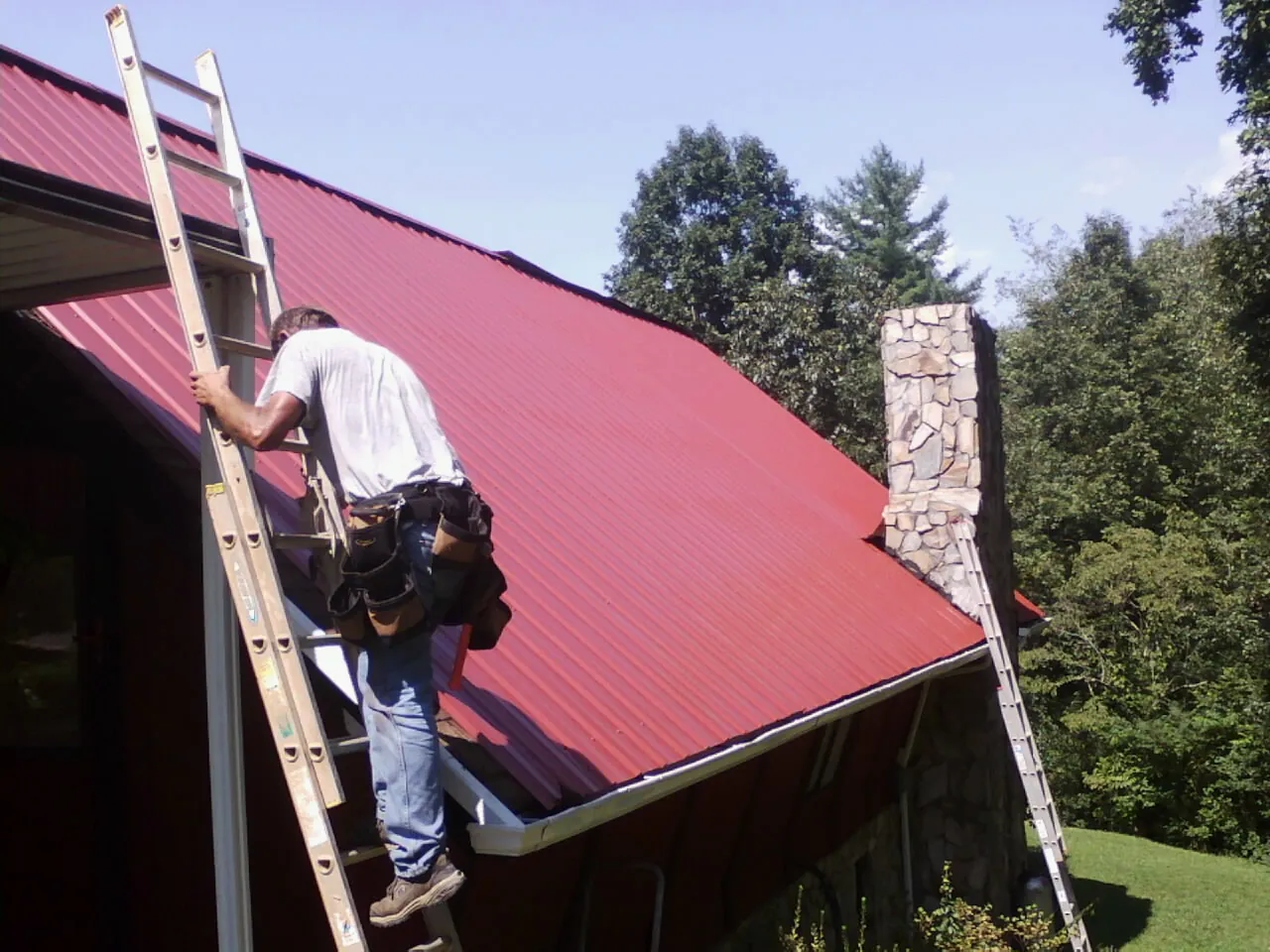 Roofer on a ladder beside a red metal roof and stone chimney — Soffit & Fascia Repair by Roofing Brothers in Waltham
